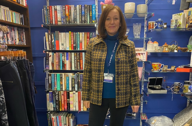 Andrea is a woman wearing a yellow checked jacket and black jeans standing in front of shelves in a charity shop.