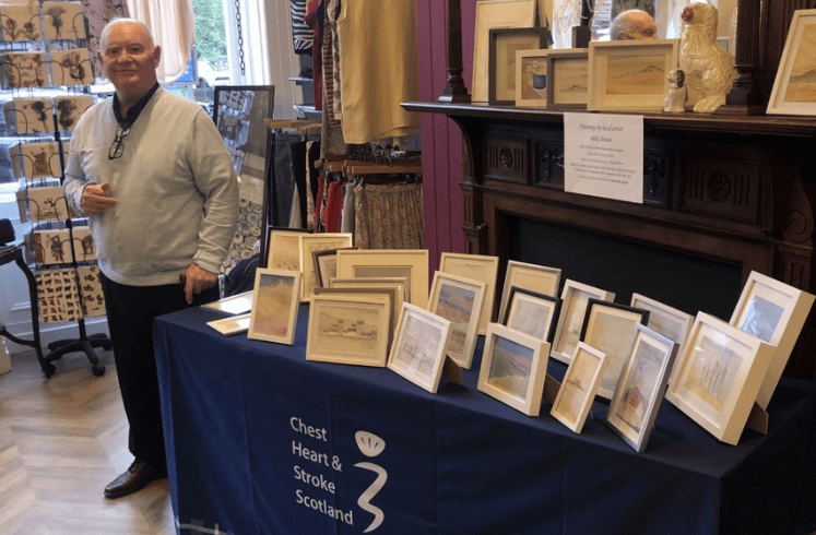 A man stands by a table displaying his framed paintings within a charity shop.