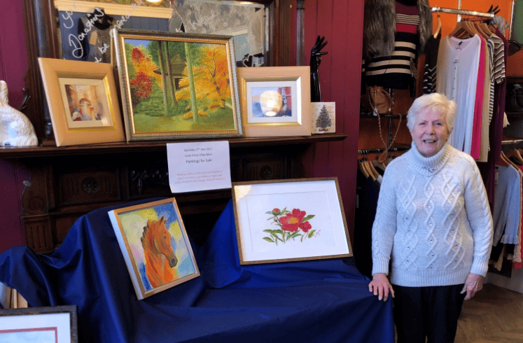 Lillias, an older woman with short grey hair, stands beside a display of her framed paintings in a charity shop.